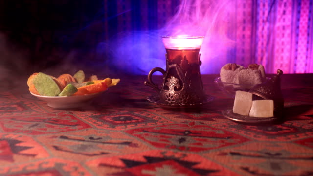 Arabischer-Tee-in-Vintage-Glas-mit-östlichen-Snacks-auf-einem-Teppich.-Östliche-Tee-Zeremonie-auf-dunklem-Hintergrund-mit-Licht-und-Rauch.-Leeren-Raum.-Selektiven-Fokus.-Schieberegler-erschossen