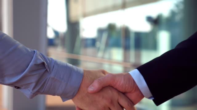 Close-up-of-two-successful-businessmen-greeting-each-other-in-urban-environment.-Young-colleagues-meeting-and-shaking-hands-near-office-building.-Handshake-of-business-partners-outdoor.-Side-view
