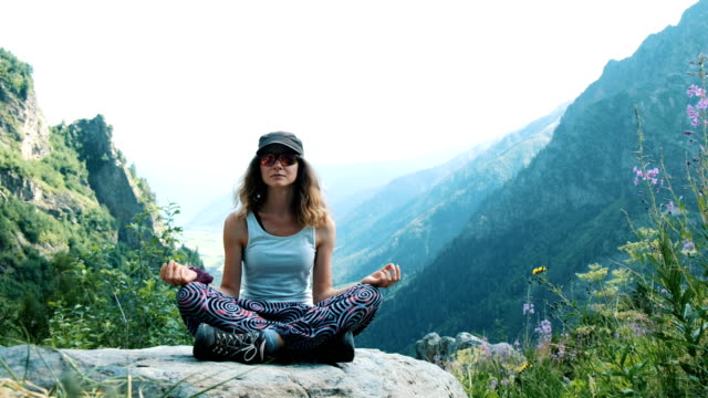 Woman-meditating-in-the-mountains-sitting-on-a-stone-in-a-lotus-pose,-a-beautiful-landscape-behind.