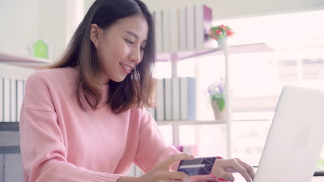 Beautiful-Asian-woman-using-computer-or-laptop-buying-online-shopping-by-credit-card-while-wear-sweater-sitting-on-desk-in-living-room-at-home.-Lifestyle-woman-at-home-concept.