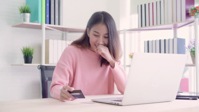 Hermosa-mujer-asiática-usando-computadora-o-laptop-comprar-online-compras-con-tarjeta-de-crédito-mientras-usar-suéter-sentada-en-el-escritorio-en-la-sala-de-estar-en-casa.-Mujer-de-estilo-de-vida-al-concepto-de-hogar.
