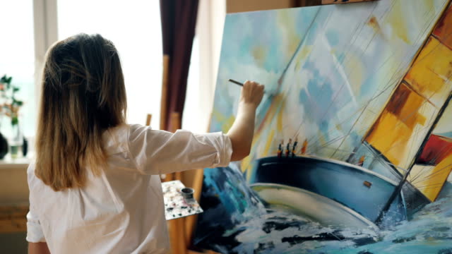 Good-looking-girl-artist-is-busy-painting-seascape-on-canvas-using-color-palette,-brush-and-canvas-on-easel-working-in-nice-studio.-Talented-people-and-work-concept.