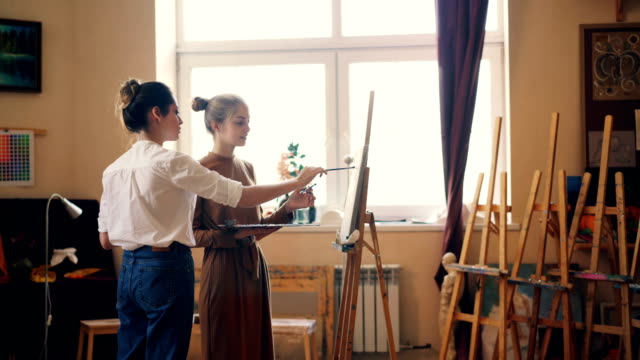 Beautiful-young-women-student-and-professional-artist-are-painting-together-in-workshop,-teacher-is-sharing-experience-and-giving-advice.-Artistry-and-creativity-concept.
