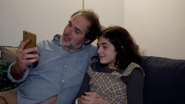 Happy-dad-and-daughter-sitting-on-couch-and-having-video-chat