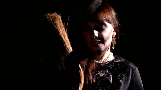 Halloween-party,-night,-frightening-portrait-of-a-woman-in-the-twilight,-in-the-rays-of-light.-woman-with-a-terrible-make-up-in-a-black-witch-costume.-she-holds-a-broom-with-a-bat
