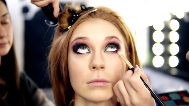 Front-footage-of-a-blonde-beautiful-model-sitting-and-looking-up-while-make-up-artist-finishing-her-smoky-eyes-.-Hairdresser-behind