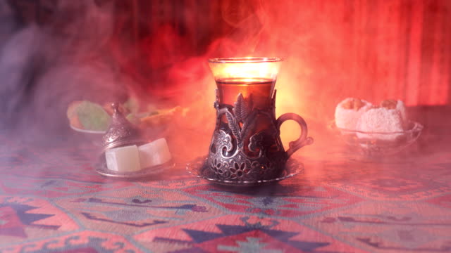 Arabian-tea-in-vintage-glass-with-eastern-snacks-on-a-carpet.-Eastern-tea-ceremony-on-dark-background-with-lights-and-smoke.-Empty-space.-Selective-focus.-Slider-shot