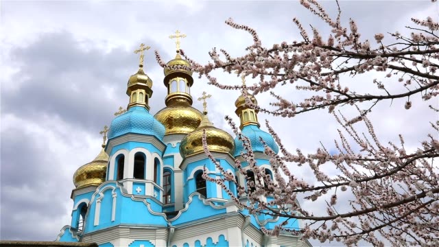 Orthodox-temple,-Clouds-above-the-temple,-golden-domes,-Timelapse,-exterior,-a-view-from-below,-View-through-the-branches-of-a-flowering-tree,-spring-landscape,-Blooming-tree