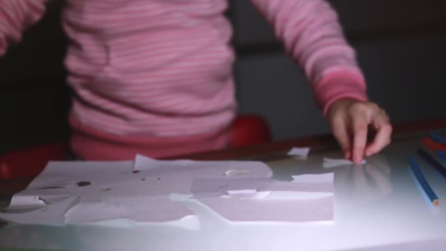 Close-up-shot-of-little-girl's-hands-in-pink-sweater-cutting-paper-sheet-shapes-with-scissors-on-a-glass-table