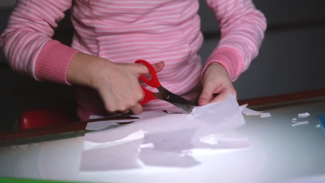 Close-up-shot-of-preschool-girl-hands-in-pink-sweater-cutting-shapes-from-paper-with-scissors-and-drawing-with-a-pencil