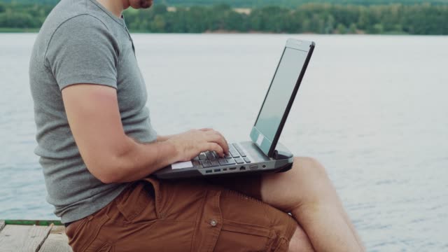 vielbeschäftigter-Mann-in-einem-grauen-T-shirt-sitzt-auf-einem-Mauerwerk-und-Texteingabe-hinter-einen-Laptop-auf-dem-Hintergrund-der-Natur