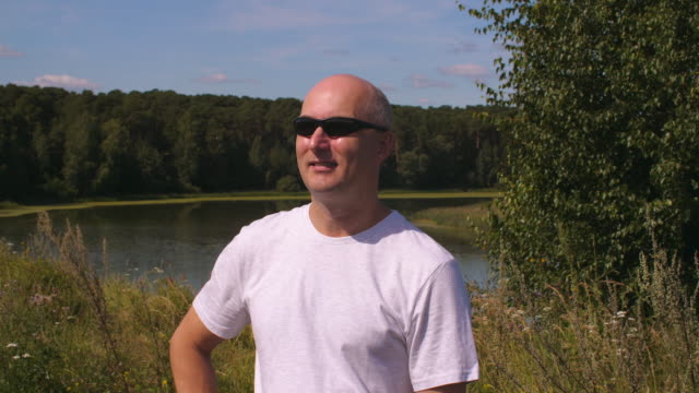 Portrait-adult-man-in-sunglasses-on-background-green-forest-and-lake-water