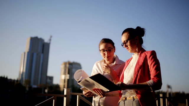 Two-women-with-the-drawings-and-technical-documentation-on-the-waterfront-on-the-background-of-skyscrapers-under-construction-to-discuss-the-risks-and-profitability-of-building-a-new-business-center.