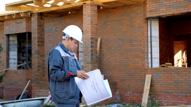 Adult-man-working-on-site-of-modern-house-and-standing-outdoors-with-paper-draft-looking-away,-slow-motion