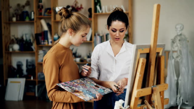Friendly-art-teacher-good-looking-woman-in-casual-clothing-is-teaching-female-student-talking-then-giving-her-brush,-girl-is-smiling-and-mixing-paints-on-palette.