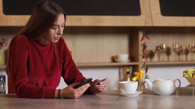 Young-woman-making-purchase-online,-typing-credit-card-details-on-black-smartphone-in-the-modern-kitchen