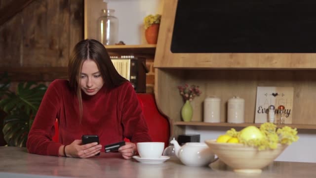 Young-woman-making-purchase-online,-typing-credit-card-details-on-black-smartphone-in-the-modern-kitchen