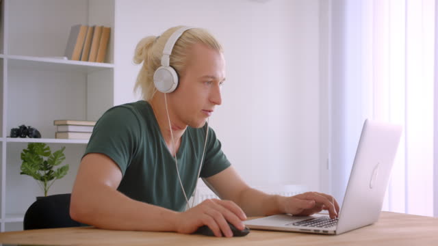 Nahaufnahme-Porträt-von-jungen-stattlichen-blonden-Hipster-Geschäftsmann-in-Kopfhörern-mit-Laptop-hören-Musik-freudig-drinnen-im-Büro