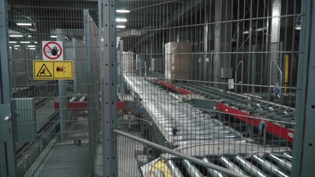 Moving-conveyor-belt-with-cardboard-boxes-along-corridor-in-workplace.-Clip.-Boxes-move-along-conveyor-belt.-Cardboard-boxes-picked-up-by-a-robot-arm-in-a-factory