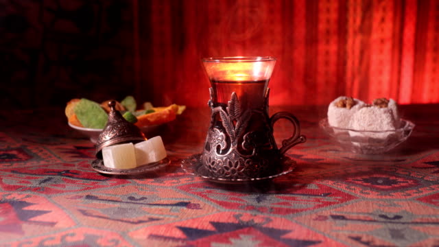 Arabischer-Tee-in-Vintage-Glas-mit-östlichen-Snacks-auf-einem-Teppich.-Östliche-Tee-Zeremonie-auf-dunklem-Hintergrund-mit-Licht-und-Rauch.-Leeren-Raum.-Selektiven-Fokus.-Schieberegler-erschossen