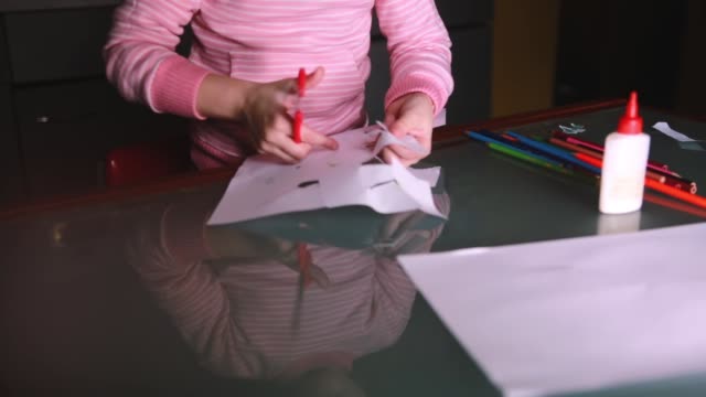 Close-up-shot-of-cute-little-girl's-hands-in-pink-sweater-cutting-shapes-from-paper,-glass-table-reflecting-her-face
