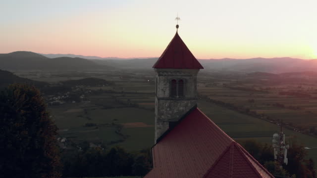 Fliegen-um-die-alte-katholische-Kirche-auf-einem-Hügel-mit-Aussicht-auf-das-Dorf-im-Sommer-in-den-Sonnenuntergang