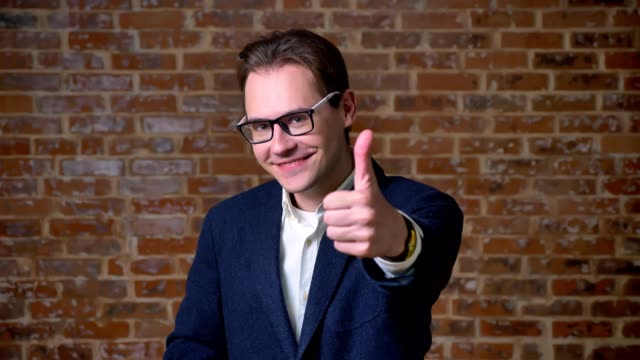 Cute-spectacled-caucasian-man-is-showing-like-sign-at-camera-and-smiling-sincerely-on-brick-background