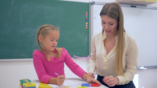 Individual-education,-Experienced-tutor-teaching-smart-girl-at-table-near-blackboard-in-classroom-of-School