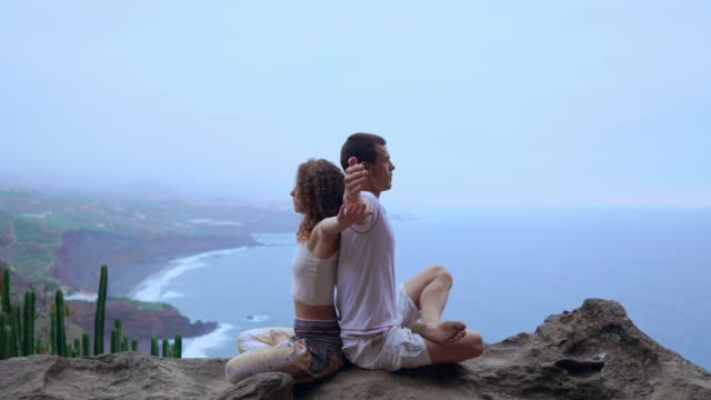 Man-and-woman-sitting-on-top-of-a-mountain-on-a-rock-back-to-back-meditate-and-do-yoga-on-the-background-of-the-ocean.