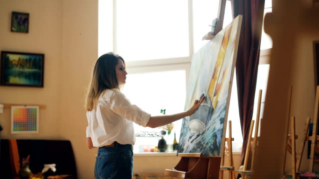Busy-young-woman-is-casual-clothing-is-painting-with-oil-paints-standing-near-easel-and-holding-brush-and-palette.-Hobby,-art-and-creative-people-concept.