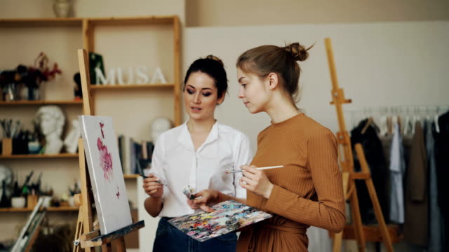 Cheerful-young-women-teacher-and-student-are-discussing-artwork-together-talking-and-looking-at-picture.-Modern-workshop-with-easels-and-tools-is-visible.