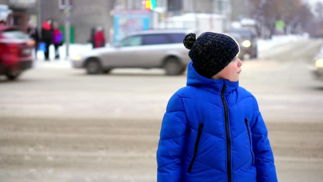 Boy-teenager-in-a-blue-down-jacket-standing-on-the-street.-Cars-are-riding-in-the-background,-the-boy-looks-around.
