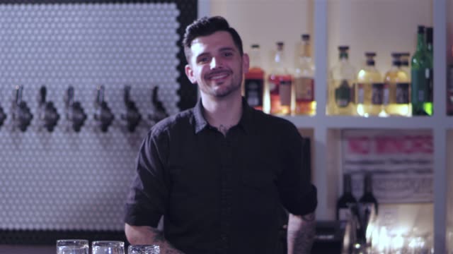 Young-attractive-male-bartender-cleaning-the-bar-with-a-towel-smiles-at-the-camera.-He-then-swings-the-towel-around-his-shoulder