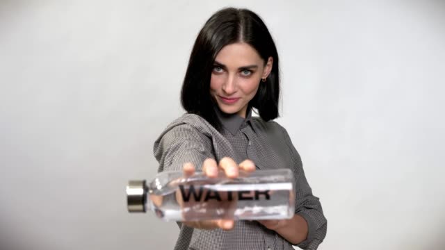 Young-pretty-woman-with-short-brown-hair-holding-and-pointing-bottle-with-water-in-camera,-showing-big-thumb,-happy,-white-background