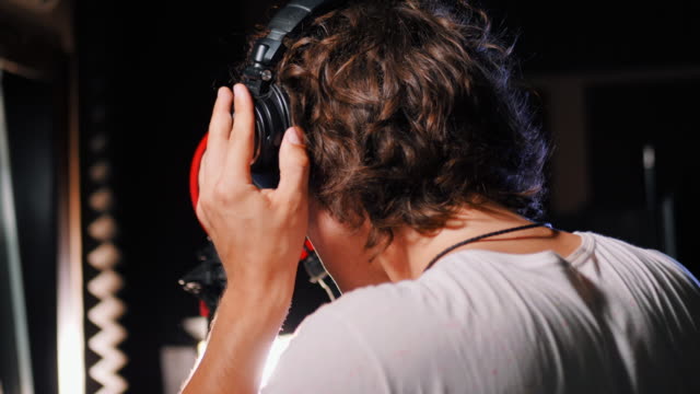 Young-handsome-singer-man-puts-on-headphones-in-the-studio.-Recording-new-melody-or-album.-Male-vocal-artist-with-curly-hair-preparing-for-working.-4k