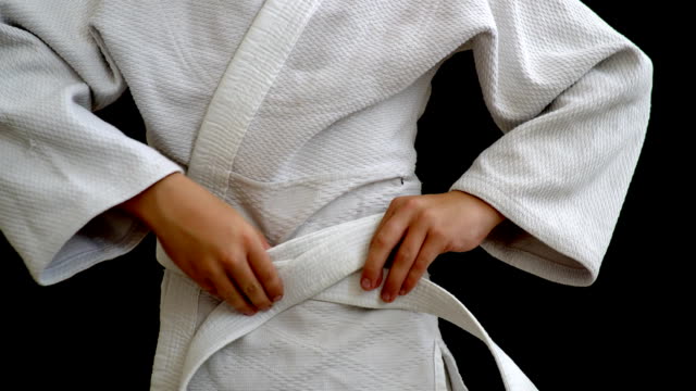 A-young-athlete-in-a-white-kimono-stands-on-a-dark-background.-The-young-man-holds-a-white-belt-in-his-hands,-his-hands-are-visible,-tying-the-belt-around-his-waist.-The-face-and-legs-are-not-visible