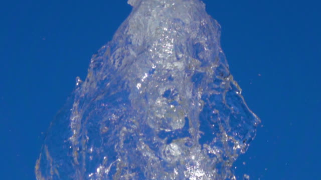 A-splash-of-clear-ice-water.-Closeup-of-light-splashing-water-in-sunny-fountain-in-park-in-summer-on-blue-sky-background.--Water-flow-of-fountain-fly-up-in-air-with-many-splashes.-Slow-motion.