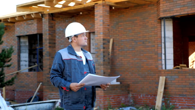 Adult-man-wearing-white-hardhat-and-looking-at-paper-plans-standing-outdoors-of-building-under-construction