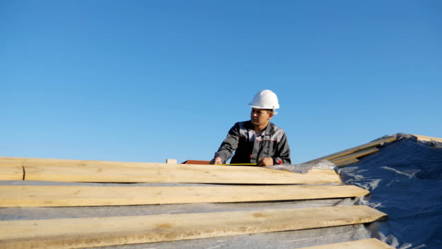 Adult-professional-builder-in-hardhat-measuring-length-of-wood-lumber-on-site-under-blue-sky