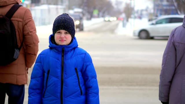 Boy-teenager-in-a-blue-down-jacket-standing-on-the-street.-Cars-are-riding-in-the-background,-the-boy-is-watching.