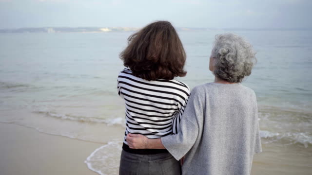 Rear-view-of-happy-senior-woman-embracing-tall-brunette-on-seashore.