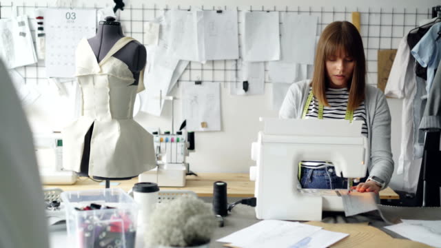 Selbstständige-Modedesigner-arbeitet-mit-Nähmaschine,-Nähen-Textile-und-Skizzen-auf-ateliertisch-liegend-zu-betrachten.-Tägliche-Arbeit-bei-Schneider-Shop-Konzept.