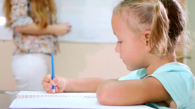 Kleine-süße-Mädchen-sitzt-im-Klassenzimmer-und-Studien-mit-Lehrer-im-Heft