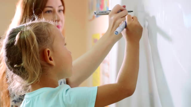 Kleine-süße-Mädchen-mit-Lehrer-schreibt-an-die-Tafel-im-Klassenzimmer