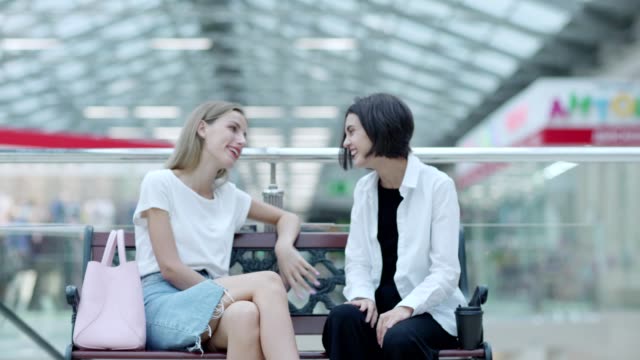Fade-in-of-two-talkative-young-women-sitting-on-bench-near-railing-in-mall-and-chatting-after-shopping