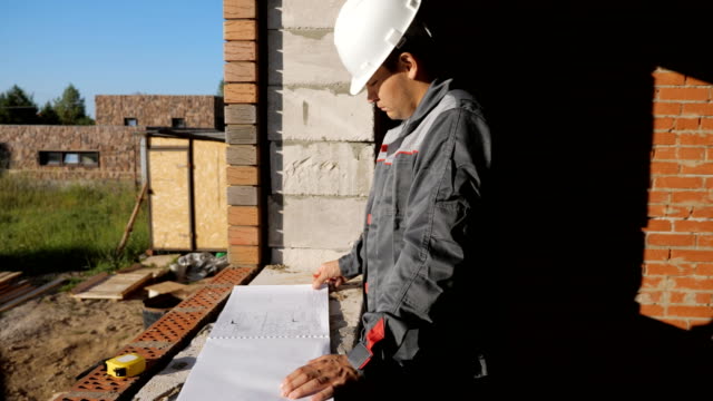 Seitenansicht-des-Mannes-in-Bauarbeiterhelm-Stand-am-Fenster-im-Haus-im-Bau-Blick-auf-Papier-Entwurf