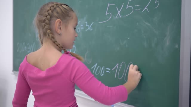 glücklich-Schulmädchen-mit-Kreide-löst-Beispiele-auf-Mathematik-auf-grüne-Schultafel-während-einer-Unterrichtsstunde-in-der-Klasse,-Grundschule