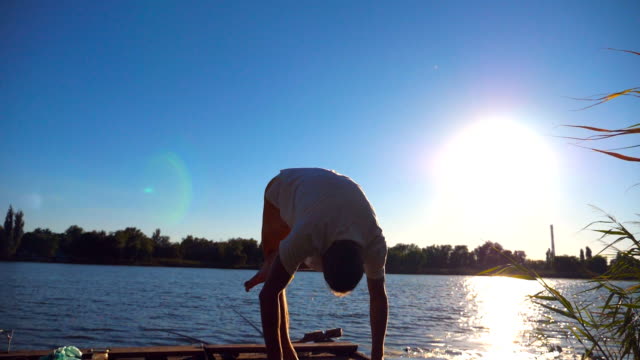 Chico-joven-practicando-yoga-se-mueve-y-coloca-en-el-borde-del-embarcadero-de-madera-en-el-lago-en-día-soleado.-Deportiva-hombre-formación-en-naturaleza-con-la-luz-del-sol-en-el-fondo.-Saludable-estilo-de-vida-activo.-Lenta-de-cerca