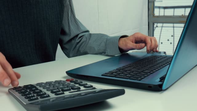 Ein-Mann-zählt-Finanzen-auf-einem-Rechner-und-gibt-Informationen-in-einen-Laptop.-Büro,-weißen-Tisch,-manager