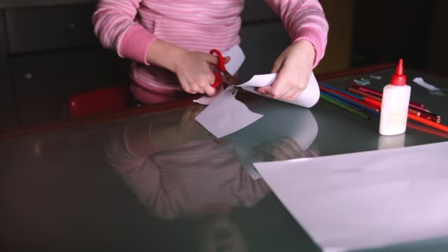 Close-up-shot-of-cute-preschool-girl-in-pink-sweater-cutting-shapes-with-scissors-from-paper,-reflecting-in-glass-table